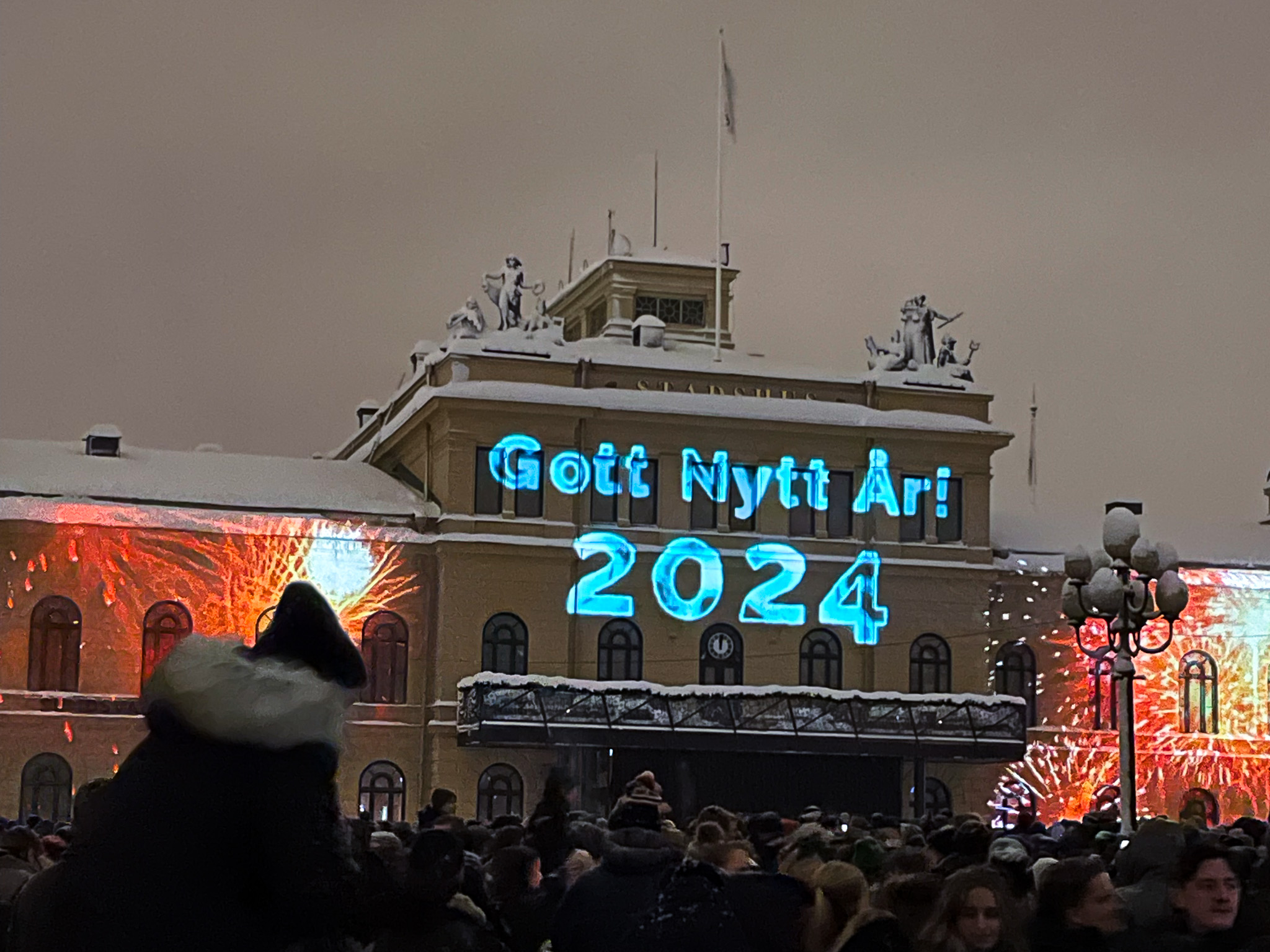 Det är många människor framför stadshuset. På Stadshusets fasad står det "Gott nytt år 2024".
